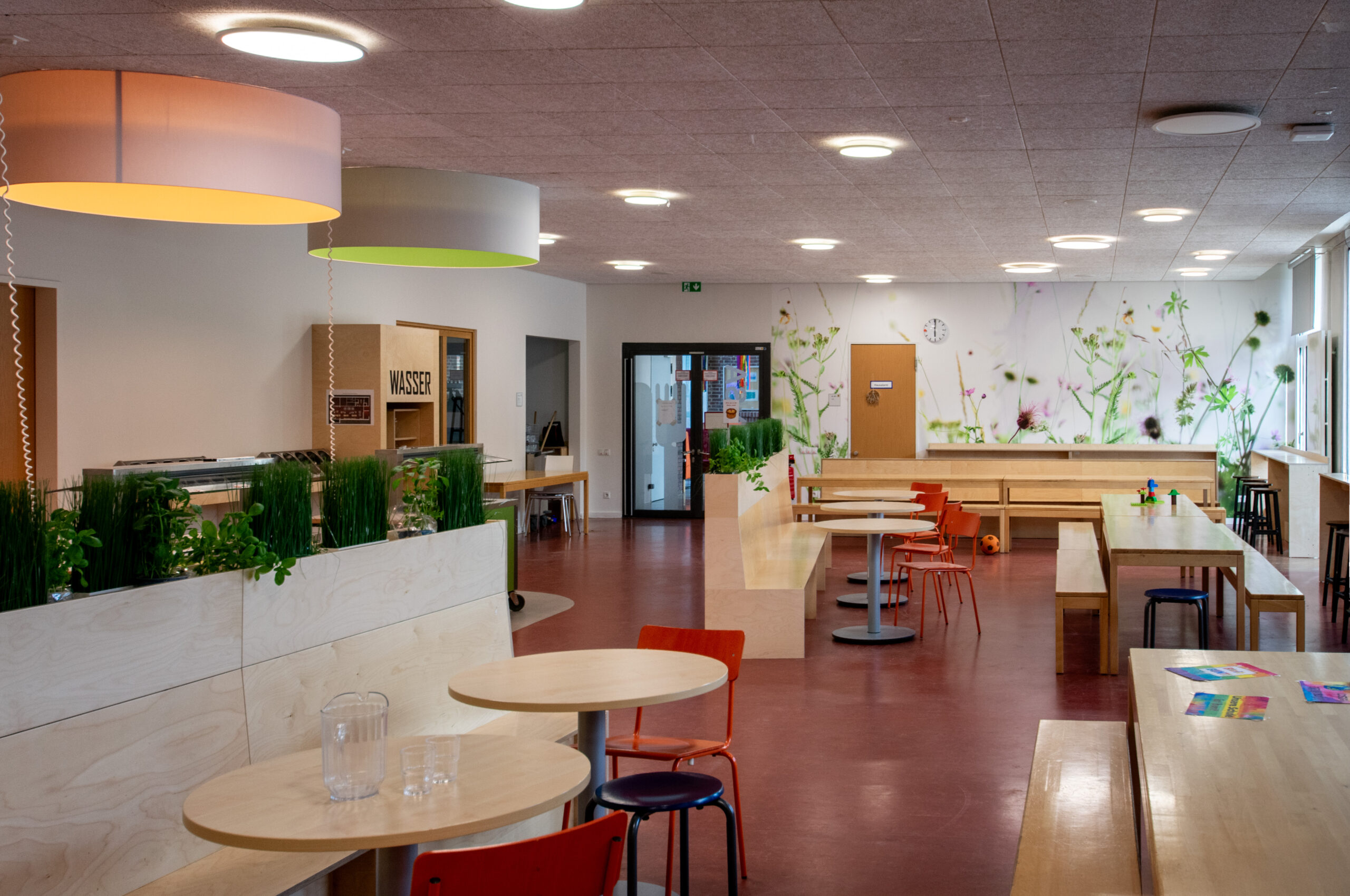 Innenansicht der von JUNISTUDIO entworfenen Mensa der Grundschule Nettelnburg mit verschiedenen Sitzbereichen, Pflanzkästen, Stehtischen und naturinspirierten Tapeten.