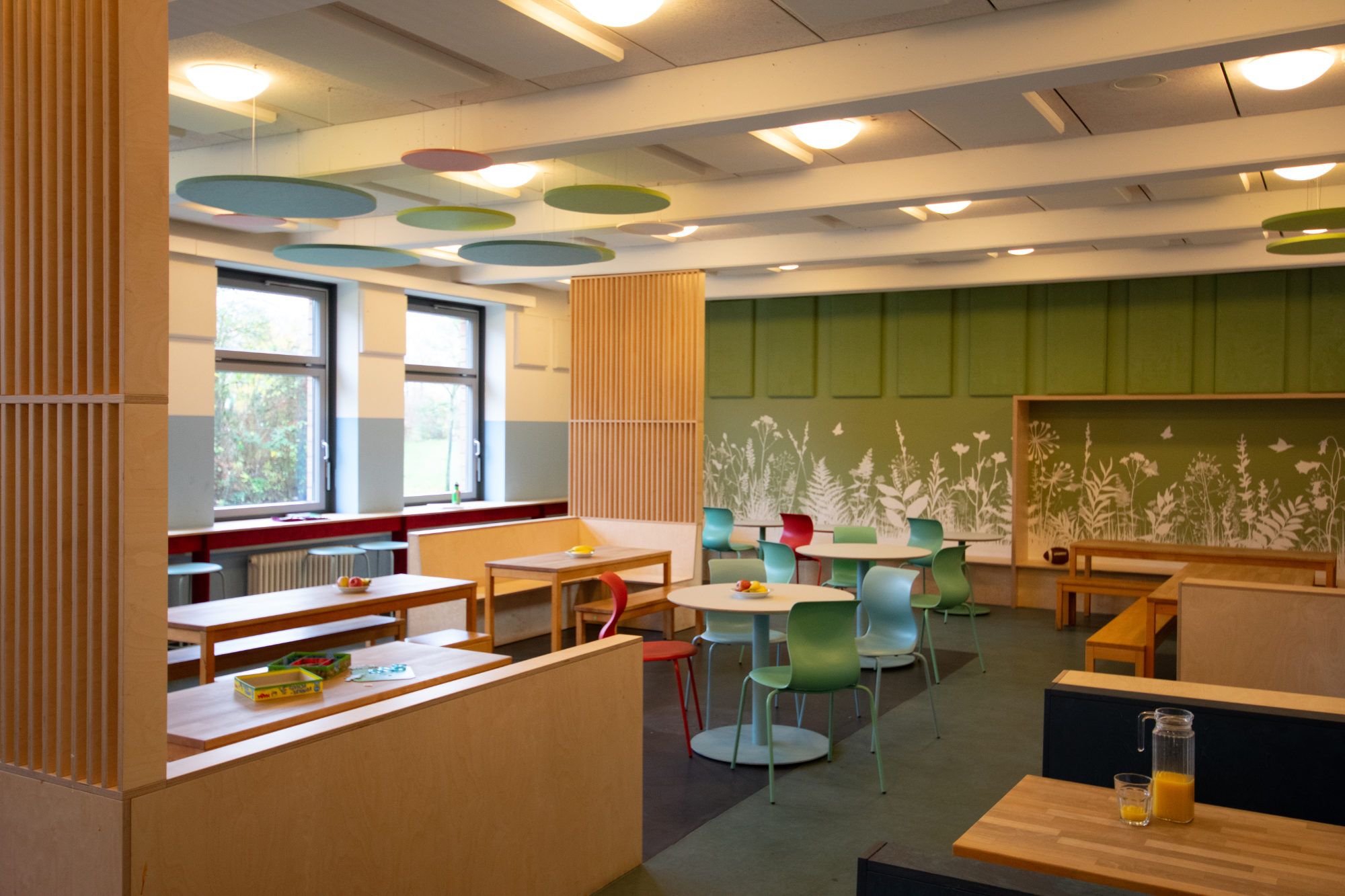 Gemütliche Sitznischen in der Mensa der Clara-Grunwald-Schule mit grüner Wandgestaltung im Naturmotiv und farblich abgestimmten Akustikpaneelen an der Decke.