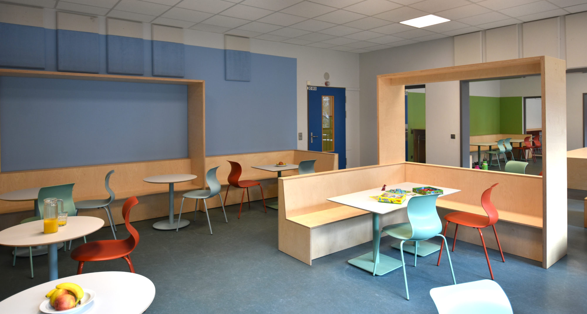Speiseraum der Clara-Grunwald-Schule mit Sitzbänken und raumbildenden Holzspangen zur Zonierung sowie runden Tischen und Stühlen in caféartiger Anordnung.