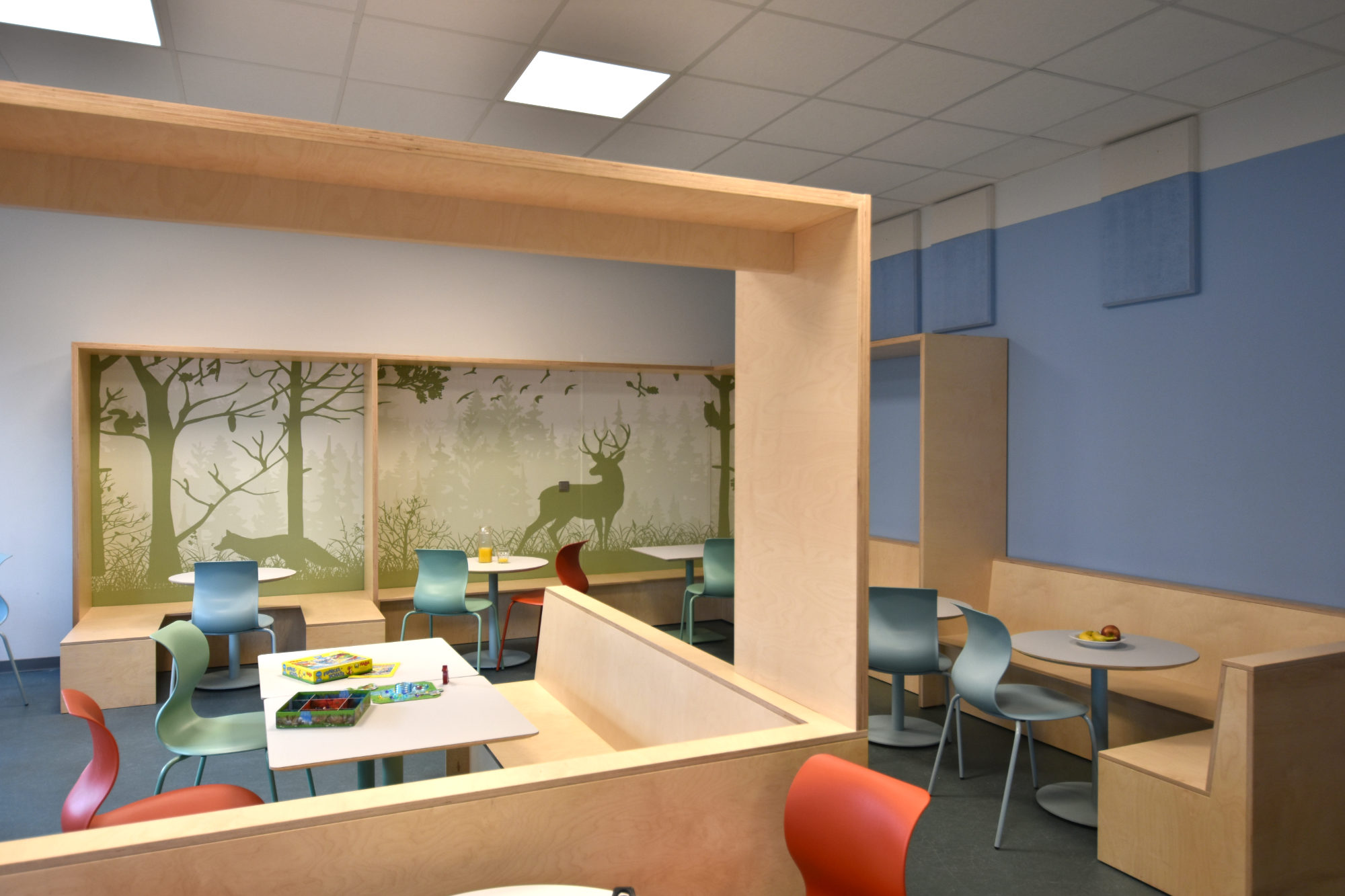 Speiseraum der Clara-Grunwald-Schule mit hölzernen Raumspangen zur Gliederung, integrierten Eckbänken und Waldmotiv-Tapete im Hintergrund.