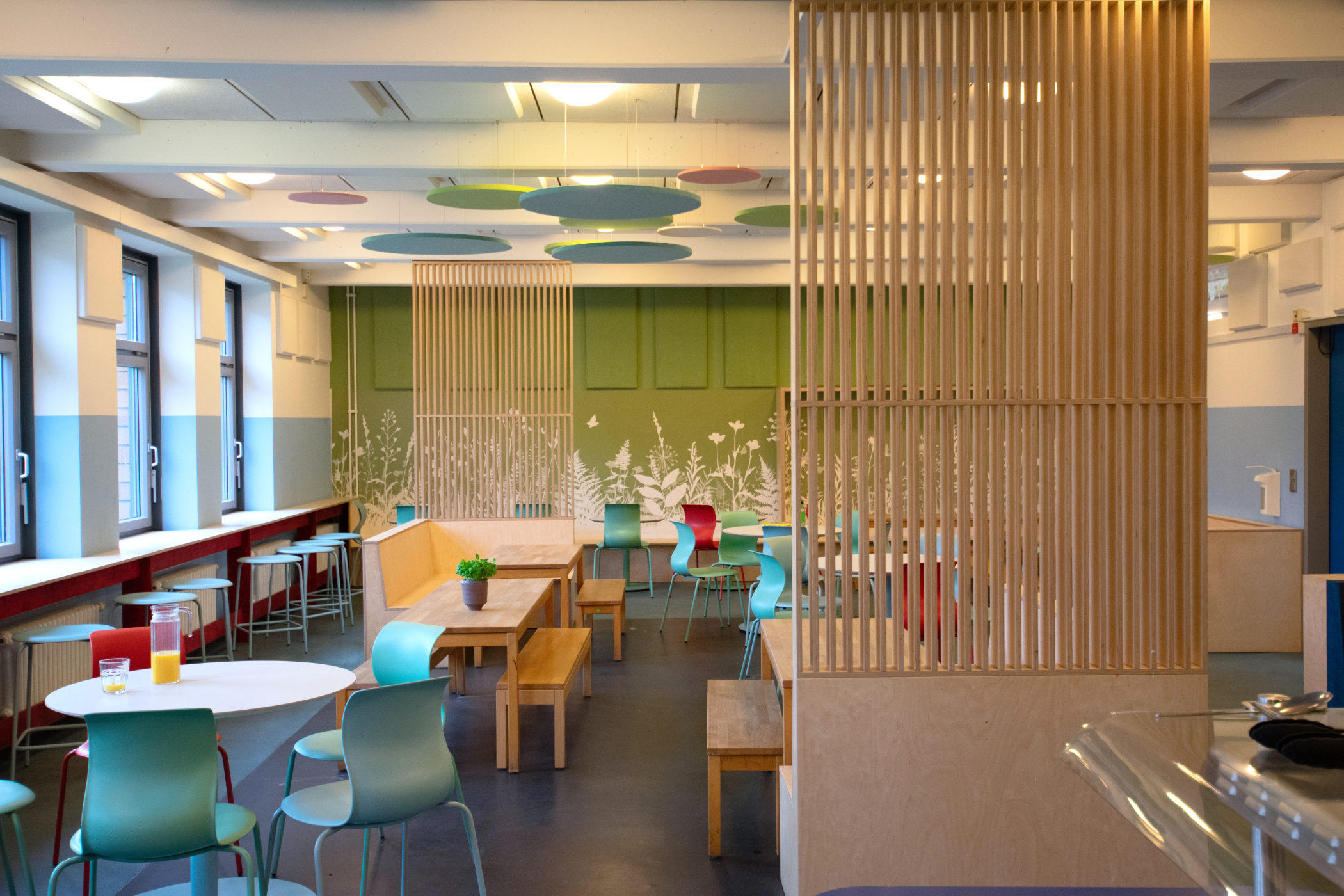 Neu gestaltete Mensa der Clara-Grunwald-Schule mit Stehtisch an der Fensterfront, Sitznischen mit Eckbänken und raumgliedernden Holzlamellen zur Zonierung.