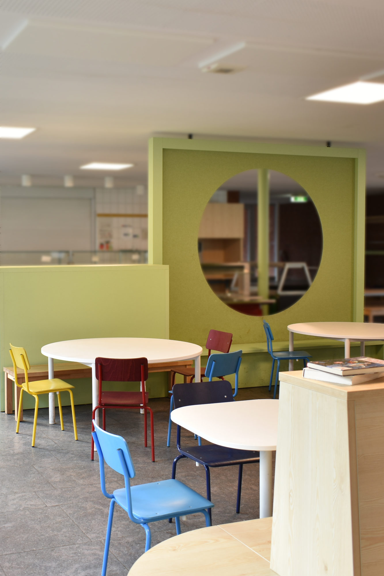 Holzsitzbank mit bunten Stühlen vor akustischem Raumteiler mit Guckloch zur Essensausgabe in einer Grundschul-Mensa