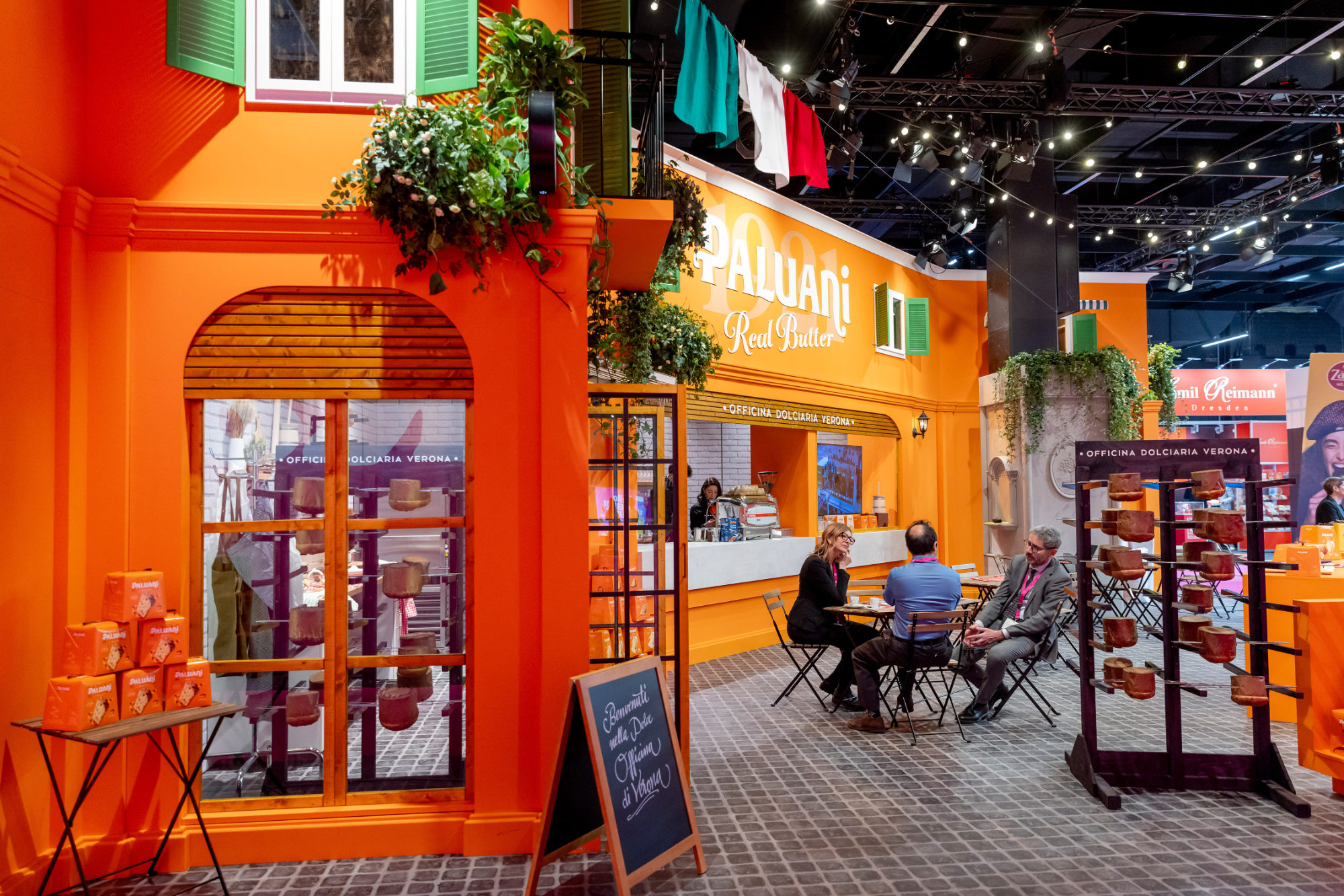 Detailansicht des von Juni Studio entworfenen Paluani Messestands auf der ISM Köln mit italienischer Piazza, Café-Sitzplätzen, Fassadenelementen und Produktpräsentation