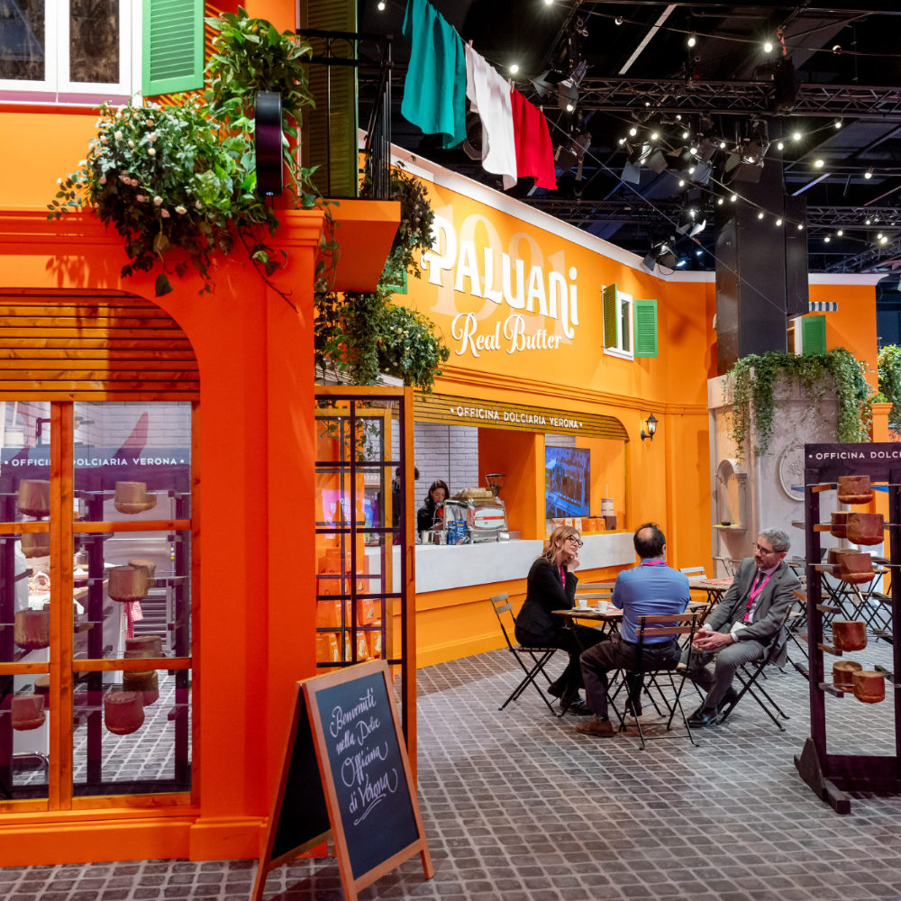 Detailansicht des von Juni Studio entworfenen Paluani Messestands auf der ISM Köln mit italienischer Piazza, Café-Sitzplätzen, Fassadenelementen und Produktpräsentation